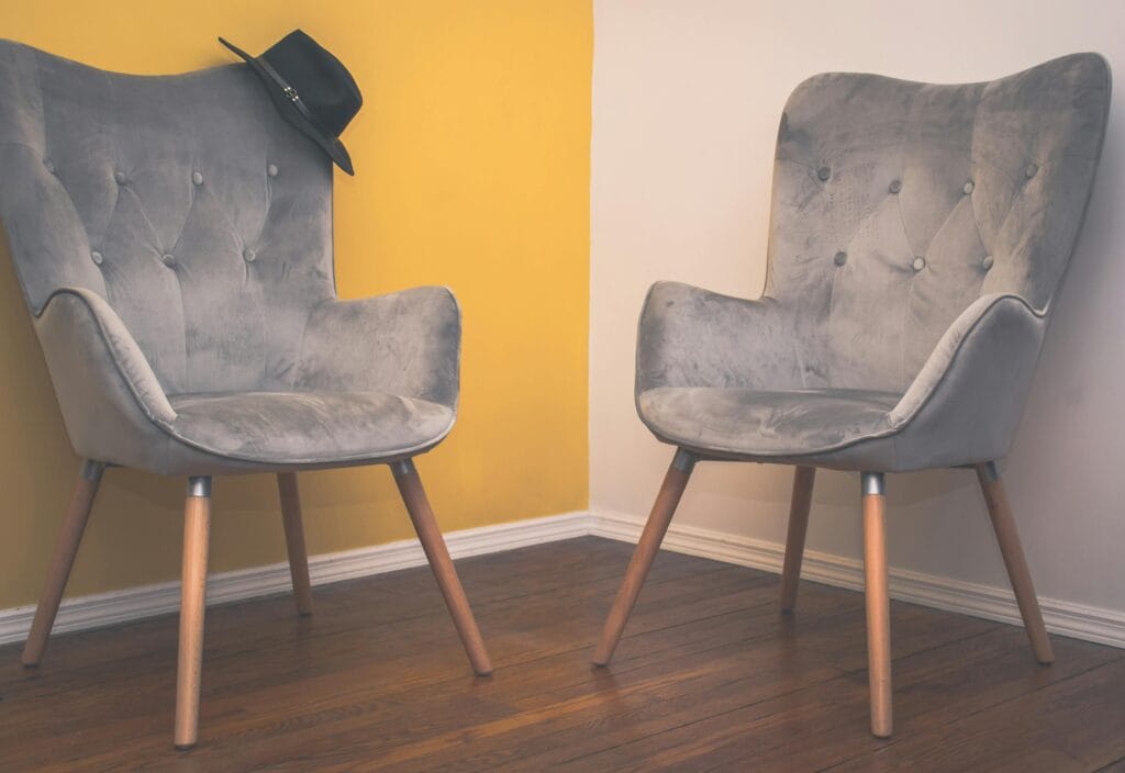 Two contemporary gray armchairs in a minimalist interior with a yellow accent wall and wooden flooring.